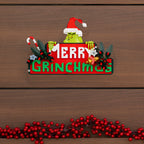 Decorative sign with Grinch face and 'Merry Grinchmas' text on a wooden background.