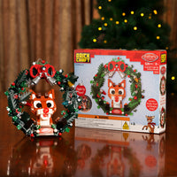 Christmas-themed figurine with wreath and packaging on a reflective surface with blurred lights in the background