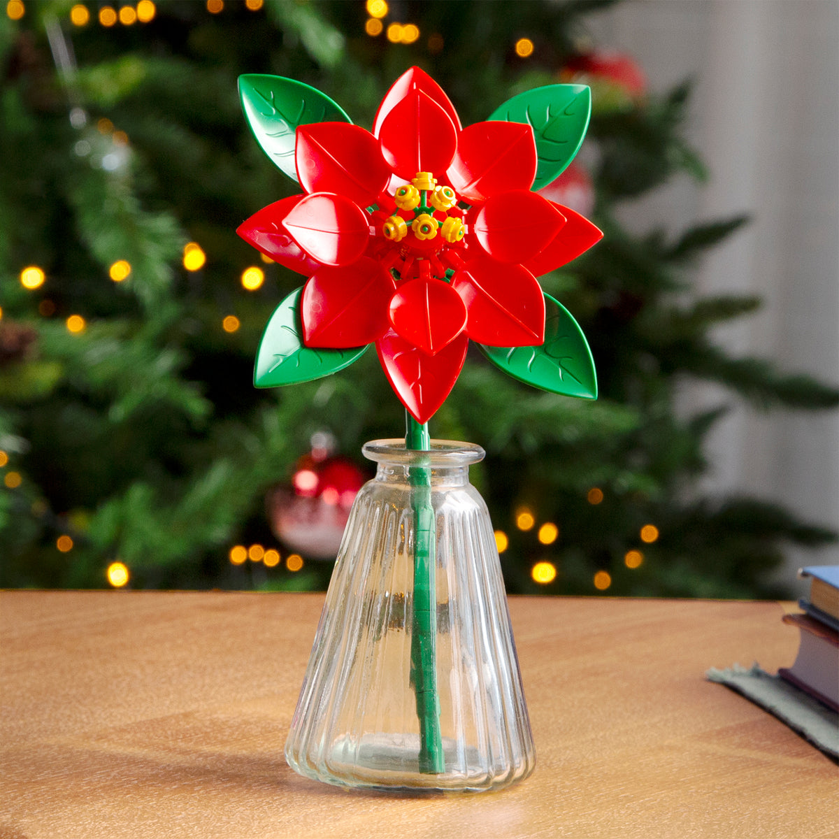 Red flower-shaped object in a clear vase on a table with a blurred Christmas tree in the background.