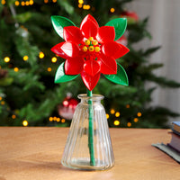 Red flower-shaped object in a clear vase on a table with a blurred Christmas tree in the background.