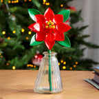 Red flower-shaped object in a clear vase on a table with a blurred Christmas tree in the background.