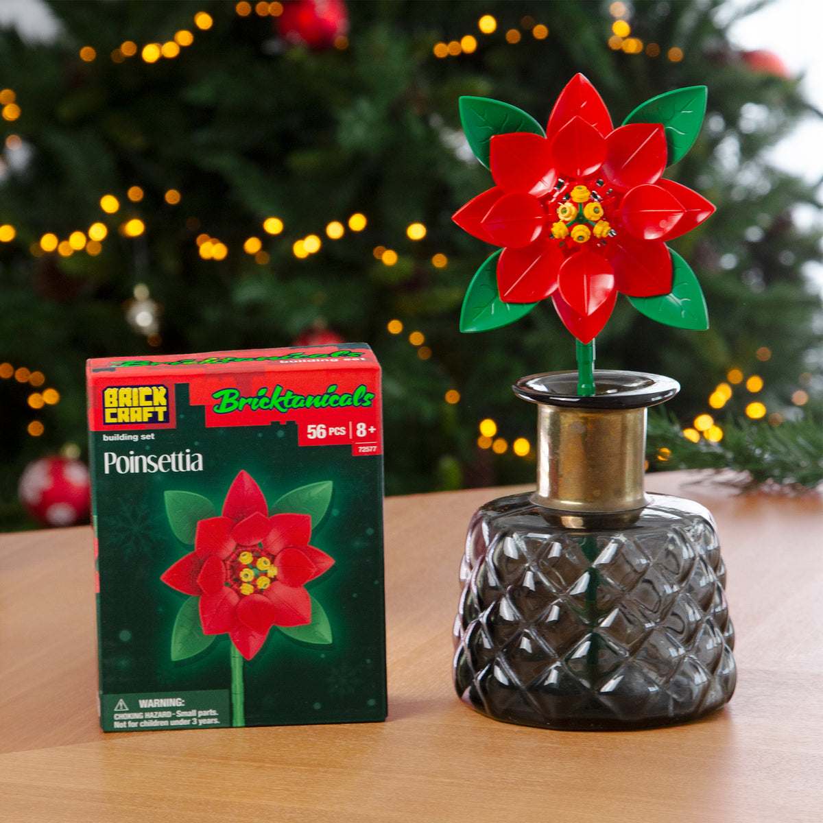 Decorative poinsettia in a vase next to its packaging in front of a Christmas tree.