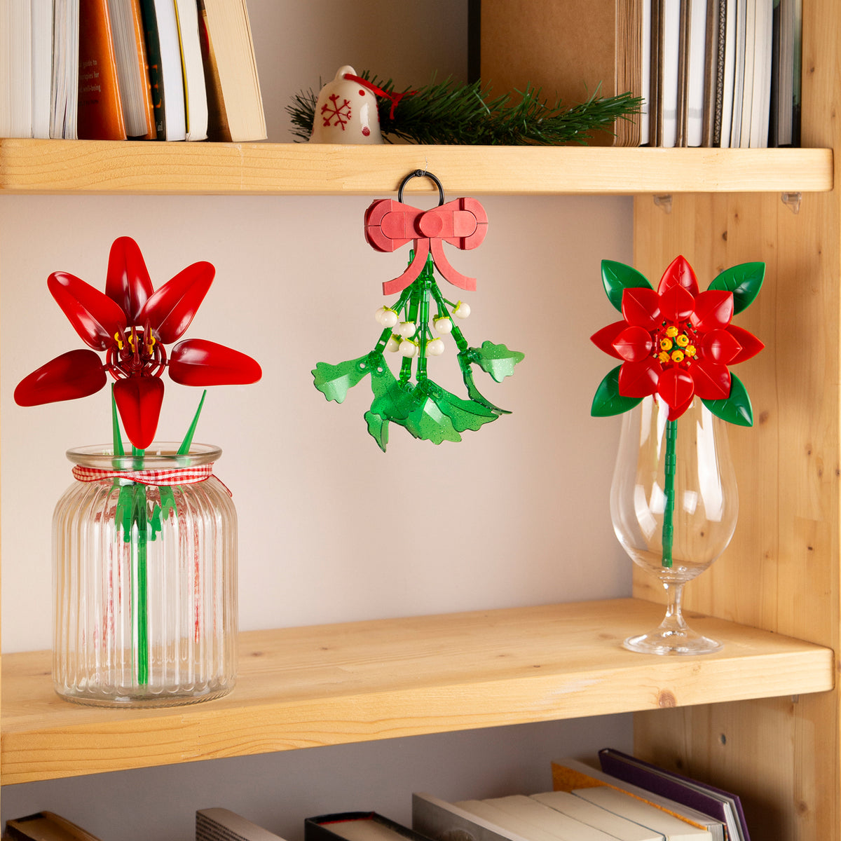 Decorative flowers in jars on a wooden shelf with books and a small Christmas tree.
