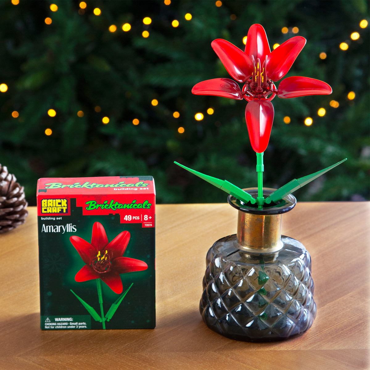Red flower in a glass vase next to its packaging on a wooden surface with a blurred festive background.